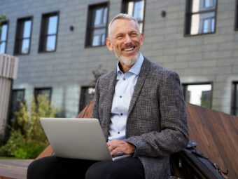 Ideas for business. Portrait of cheerful elegant middle aged businessman smiling while working on his laptop, sitting on the bench near modern building outdoors