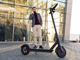 Young handsome man with e-scooter standing on sidewalk near the airport on the sunset and talking on the smartphone.Trendy urban transportation on modern electric scooter.Eco friendly mobility concep
